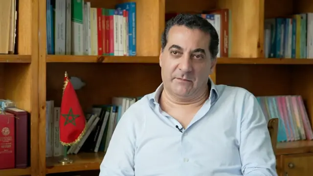 رجل يجلس مع مكتبة الكتب والعلم المغربي خلفه